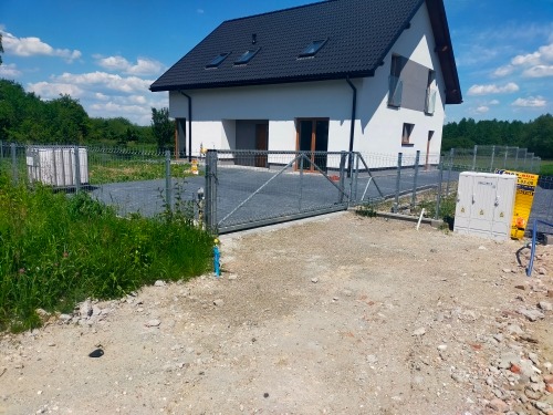 Nowoczesny dom jednorodzinny z czarnym dachem i szarą bramą wjazdową, widoczne skrzynki elektryczne przed posesją.