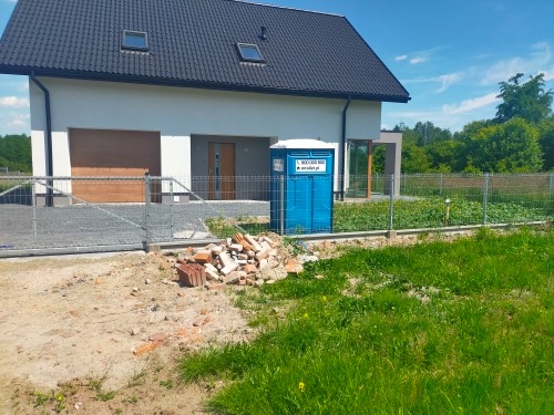 Nowoczesny dom jednorodzinny z ciemnym dachem, widoczna niebieska toaleta przenośna na posesji oraz sterta cegieł na pierwszym planie.