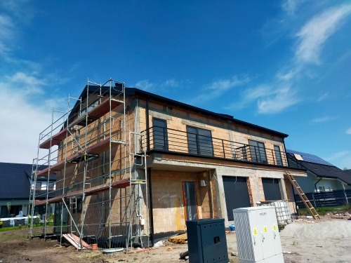 Dwukondygnacyjny dom murowany w trakcie budowy z widocznymi rusztowaniami, niezadaszonym balkonem z czarną balustradą, oknami w czarnych ramach, z elementami instalacji elektrycznej na zewnątrz...