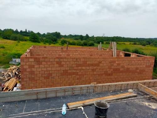 Wznoszone mury domu z bloczków ceramicznych na tle zieleni, widoczne zbrojenie fundamentów i butelka wody na dachu.