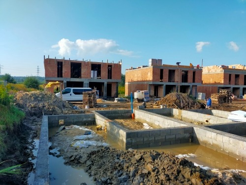 Budowa bliźniaczych domów w stanie surowym otwartym: widoczne fundamenty zalane wodą, ściany parteru z czerwonej cegły, częściowo wzniesione ściany piętra, biały samochód dostawczy w tle.