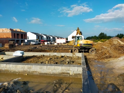 Fundamenty nowego osiedla w trakcie budowy, widoczna żółta koparka Liebherr i rozpoczęte budynki z cegły oraz gotowe domy w tle pod błękitnym niebem.