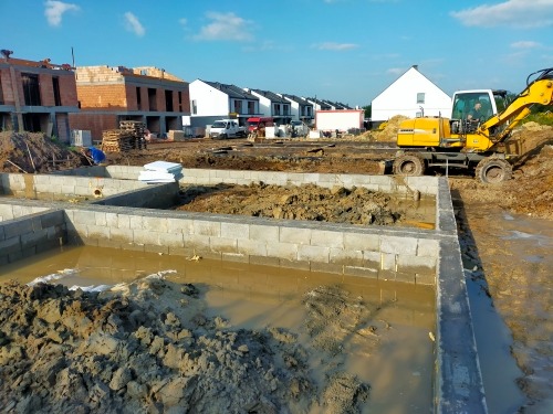 Fundamenty murowanego domu w trakcie budowy, wypełnione wodą, z widoczną koparką i budynkami w tle pod błękitnym niebem.