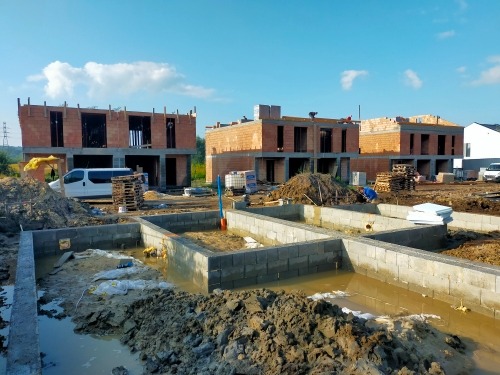 Fundamenty pod budowę trzech domów jednorodzinnych z betonu komórkowego, widoczne częściowo wzniesione ściany, palety z materiałami budowlanymi i zaparkowany bus w tle.