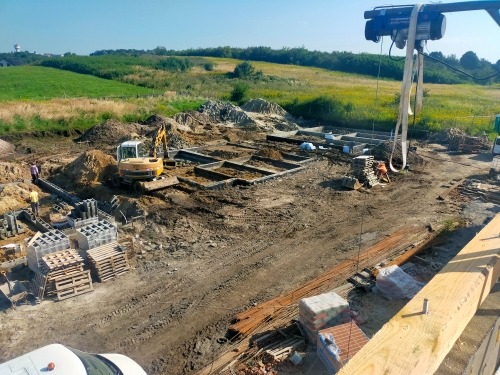 Fundamenty domu w trakcie budowy: wykopane rowy, szalunki, zbrojenie, palety z bloczkami betonowymi, mała koparka na placu budowy, widok na zielone pola w tle.