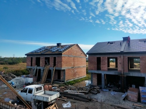 Dwa domy w stanie surowym otwartym, jeden z panelami solarnymi na dachu, drugi z ułożoną dachówką, na dachu którego stoi dekarz w czerwonym kombinezonie, na pierwszym planie biały samochód...