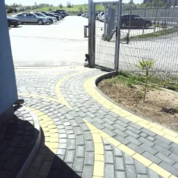Ścieżka z szarej kostki brukowej z żółtymi akcentami prowadząca od progu budynku do bramy wjazdowej na parking z widocznymi samochodami w tle.