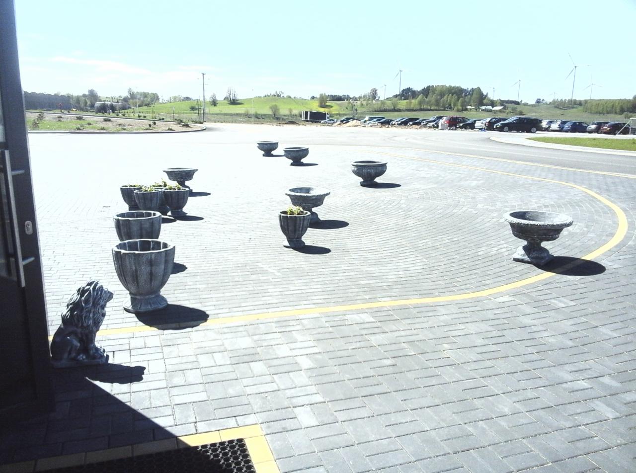 Szara kostka brukowa ułożona w łuk z żółtą linią, ozdobne betonowe donice wzdłuż drogi, figura lwa przy wejściu do budynku, parking z samochodami i turbiny wiatrowe w tle.