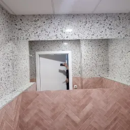 Wykończona łazienka z lustrem, ściany wyłożone płytkami imitującymi lastryko w kolorze szaro-białym, dolna część ścian wyłożona płytkami w odcieniach różu ułożonymi w jodełkę. W lustrze odbija się...