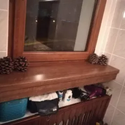 Brązowy parapet z drewna pod oknem w łazience, ozdobiony szyszkami, z widoczną przestrzenią pod spodem, gdzie znajdują się różne przedmioty, w tym suszarka do włosów.