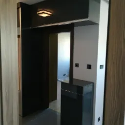 Ciemna zabudowa szafkowa nad wejściem do pokoju, z dodatkową szafką stojącą, oświetlona lampą sufitową, widok z korytarza.