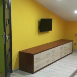 Długa komoda z szufladami w dwukolorowej obudowie, umieszczona pod telewizorem na żółtej ścianie w pokoju na poddaszu.