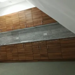 Komplet szafek kuchennych z drewnianymi frontami i szarym blatem, zamontowany w pomieszczeniu ze skośnym sufitem i szarą glazurą na ścianie.