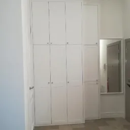 Wysoka, biała szafa wnękowa z czterema rzędami drzwi, dopasowana do sufitu, z widocznym lustrem i fragmentem przedpokoju.