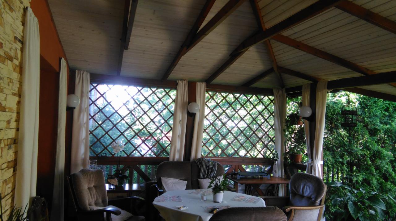 Zadaszona weranda z drewnianą konstrukcją, wyposażona w meble wypoczynkowe, stół nakryty obrusem oraz okna z kratownicą i zasłonami.