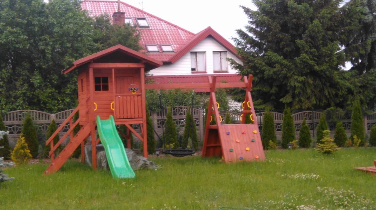 Drewniany plac zabaw z domkiem, zjeżdżalnią i ścianką wspinaczkową na tle domu z czerwoną dachówką i zielonego trawnika.