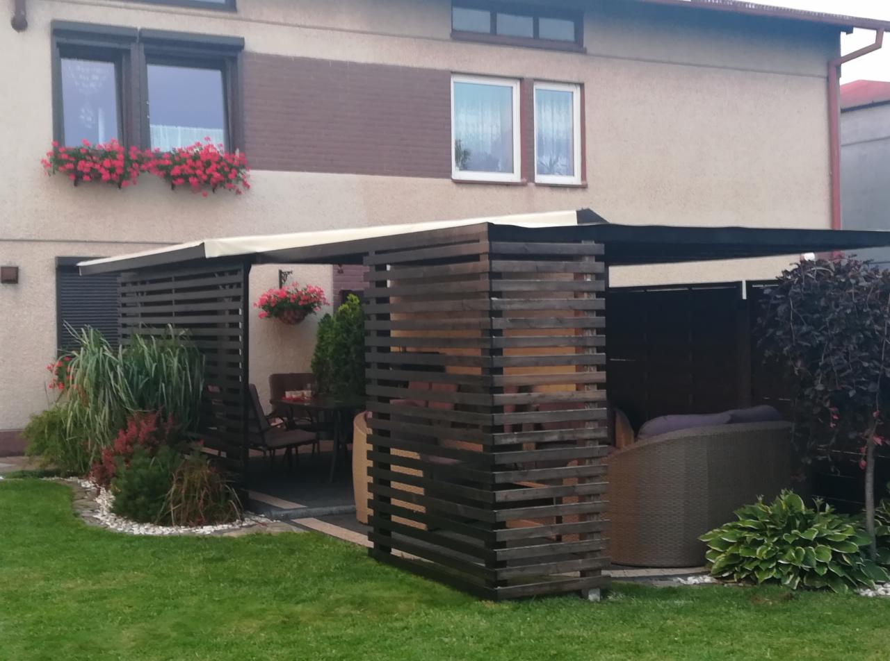 Drewniana pergola z lamelami w ogrodzie, zadaszona, z meblami ogrodowymi w środku, kwiaty w donicach wiszących i na parapecie okna
