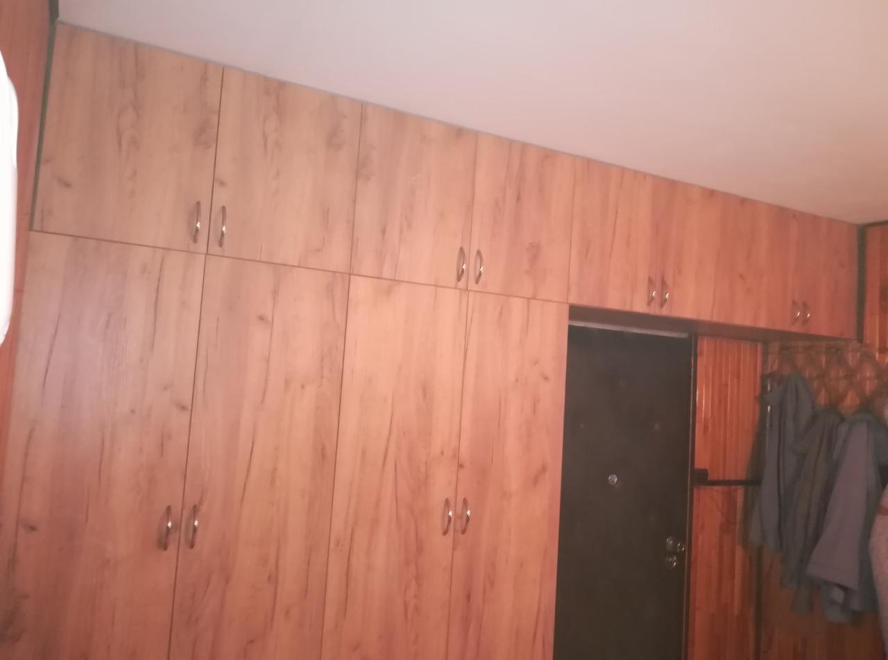 Zabudowa szafy z drewnopodobnymi frontami, sięgająca sufitu, z widocznymi uchwytami i drzwiami wejściowymi w tle.