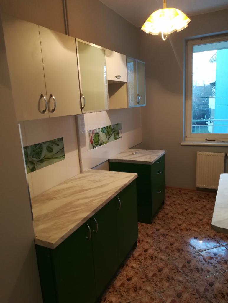 Wykończona kuchnia z zielonymi szafkami dolnymi i białymi górnymi, blatem imitującym marmur i płytkami z motywem roślinnym na ścianie, oświetlona lampą sufitową. Widoczny fragment okna z widokiem...