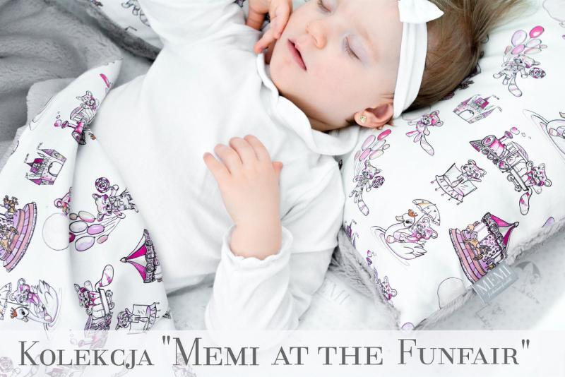 Śpiące niemowlę w białym ubranku, leżące na poduszce i otulone kocykiem z motywem wesołego miasteczka w odcieniach fioletu i różu. Widoczny złoty kolczyk w uchu dziecka.