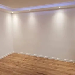 Pusty pokój po remoncie z panelami podłogowymi w odcieniach brązu, świeżo pomalowanymi białymi ścianami i oświetleniem LED w suficie podwieszanym.