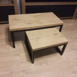 Dwa stoliki kawowe o drewnianych blatach i metalowych, czarnych nogach, ustawione jeden pod drugim, na podłodze z paneli imitujących drewno.