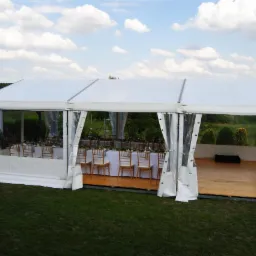 Biały namiot imprezowy z drewnianą podłogą i dekoracją kwiatową, widok na zewnątrz z nakrytymi stołami i krzesłami, ustawiony na trawie pod błękitnym niebem z chmurami.