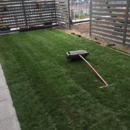 Świeżo ułożona trawa z rolki na małym trawniku, widoczny walec do trawy, fragmenty rolek trawy przy ogrodzeniu.