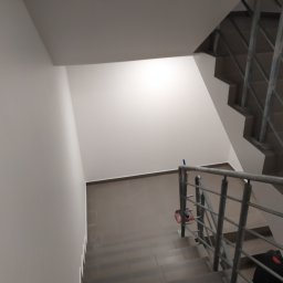 RESIN MADE ART - Klatka schodowa wykończona brązowymi płytkami, widok z góry na dół, z widoczną balustradą ze stali nierdzewnej i fragmentem ściany pomalowanej na biało. Na jednym z stopni leży czerwona szmatka.