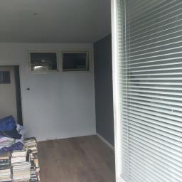 RESIN MADE ART - Pokój po remoncie z nową, jasną podłogą imitującą drewno, sterta książek pod ścianą, szare ściany i rolety w oknie.