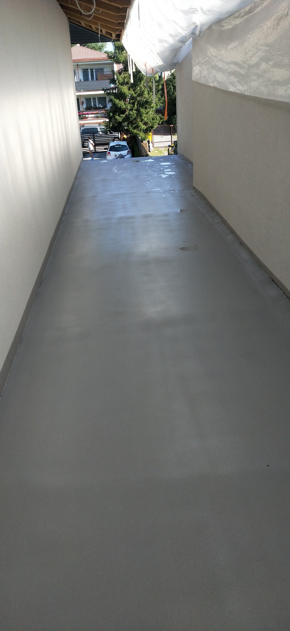 Świeżo wylana, szara posadzka w wąskim przejściu między budynkami, z widocznymi odbiciami światła i wilgoci na powierzchni. W tle widoczny budynek mieszkalny i zieleń drzew.