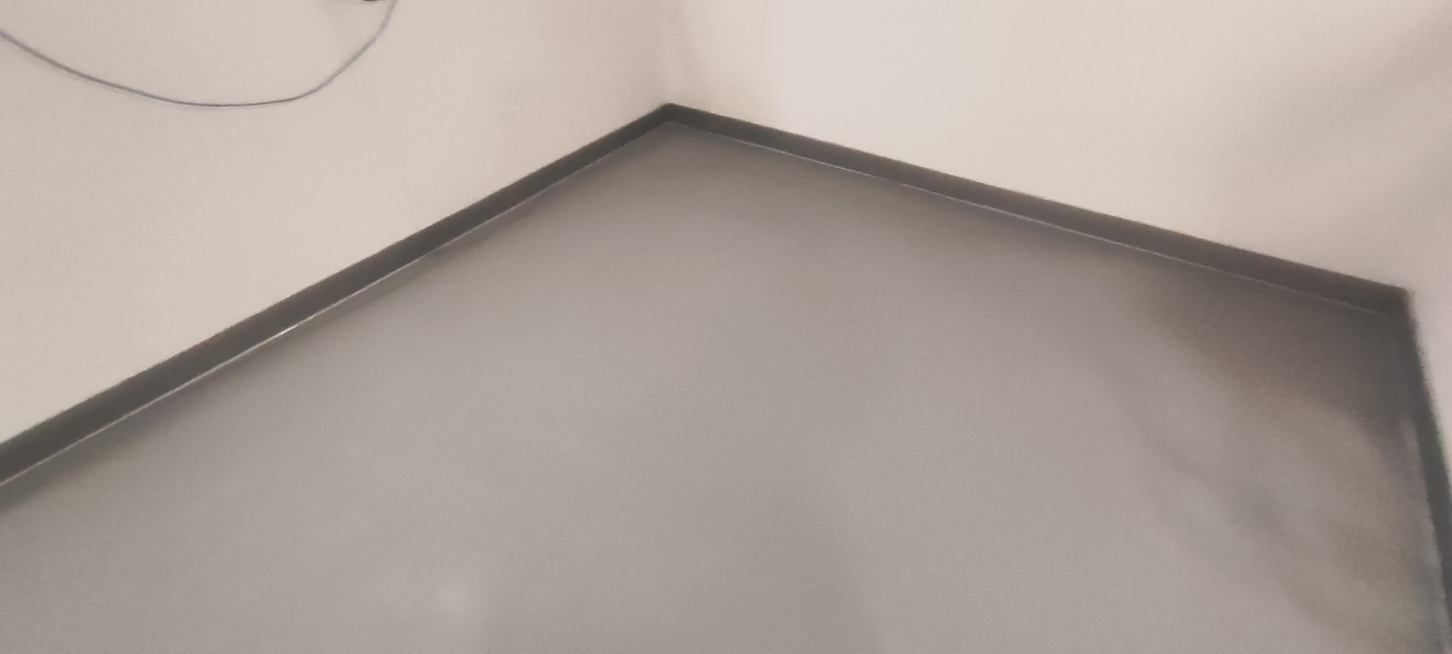 Gładka, szara posadzka w rogu pomieszczenia, widoczne listwy przypodłogowe i fragment kabla na ścianie.