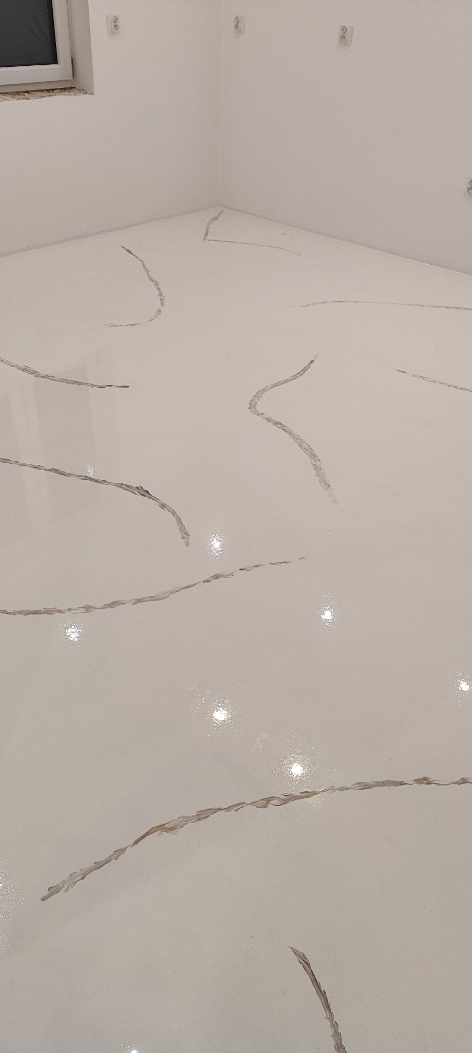 Biała posadzka żywiczna z ciemniejszymi, nieregularnymi wzorami przypominającymi marmur, widoczna w rogu pomieszczenia z białymi ścianami i gniazdkami elektrycznymi.