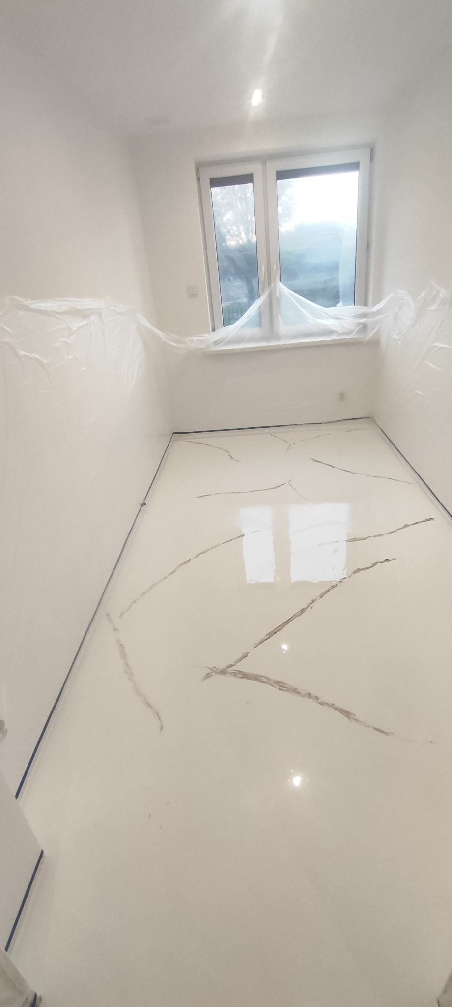 Jasne wnętrze z nową, błyszczącą podłogą z wzorem imitującym marmur, zabezpieczone folią ściany i okno.