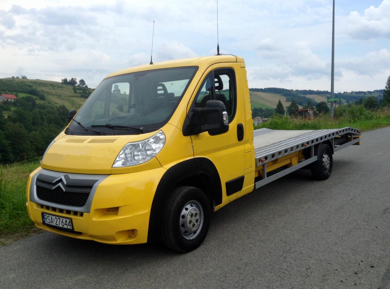 Żółty samochód laweta marki Citroen na asfaltowej drodze, w tle pagórkowaty krajobraz z zielenią i zabudowaniami.