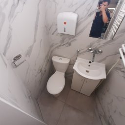 MMBUD - Nowoczesna, wykończona łazienka z marmurowymi płytkami ściennymi, białą umywalką z szafką, toaletą i podajnikiem na papier, uchwycona w odbiciu lustrzanym z widoczną osobą robiącą zdjęcie telefonem.