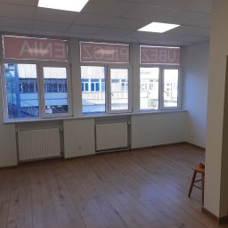 MMBUD - Puste biuro z drewnianą podłogą, białymi ścianami i grzejnikami pod oknami z napisem na szybach widzianym od wewnątrz, mały drewniany stołek w rogu.