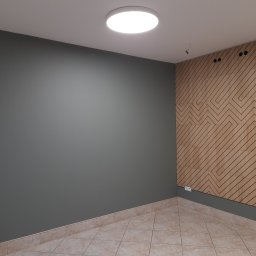 MMBUD - Szara ściana pomalowana farbą w pokoju z drewnianą ścianą dekoracyjną z wzorem zygzaków, jasna podłoga z płytek i okrągła lampa sufitowa.