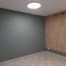 Szara ściana pomalowana farbą w pokoju z drewnianą ścianą dekoracyjną z wzorem zygzaków, jasna podłoga z płytek i okrągła lampa sufitowa.