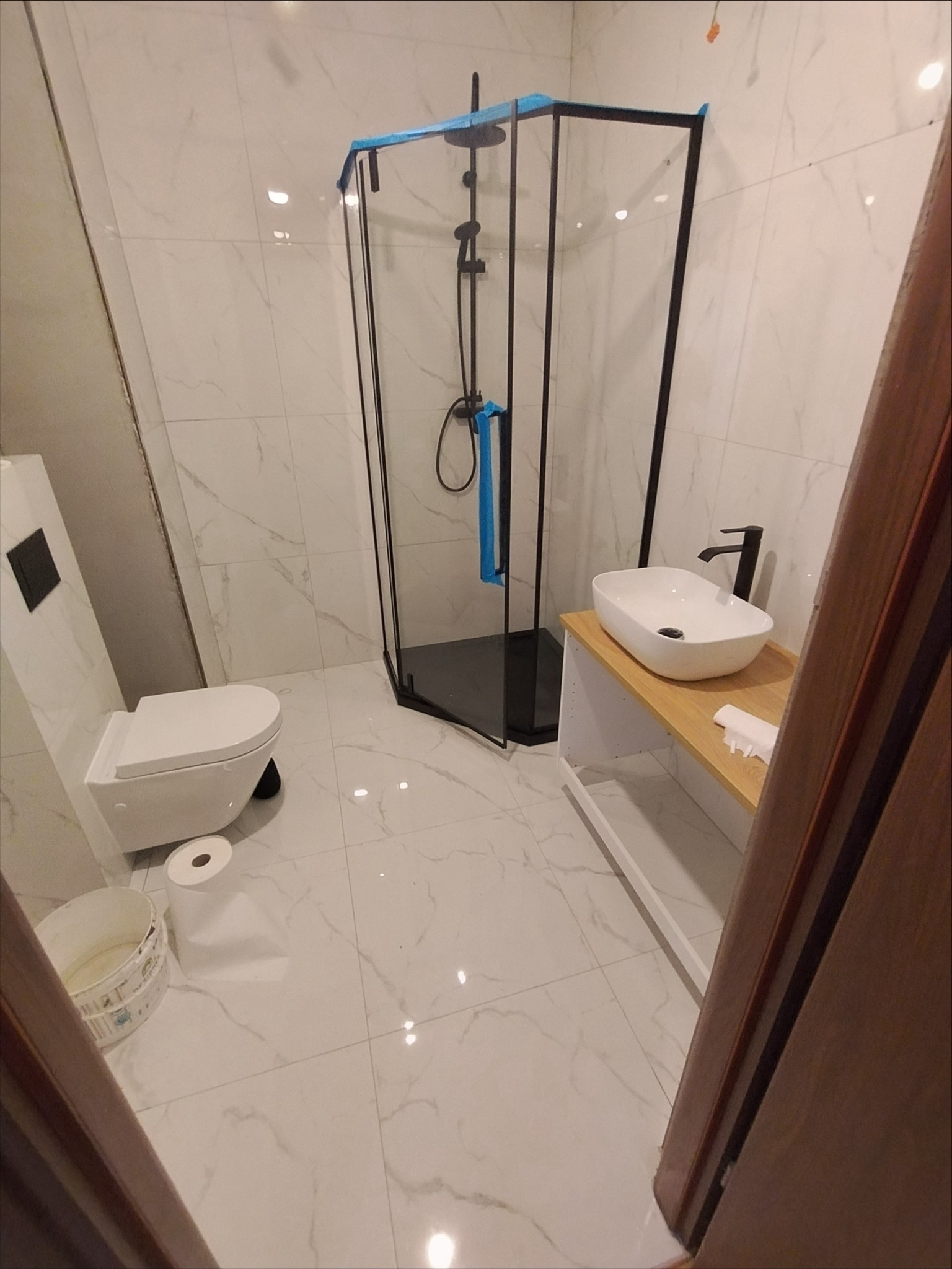 Nowoczesna łazienka z kabiną prysznicową typu walk-in z czarnymi profilami, wiszącą toaletą i umywalką nablatową na drewnianym blacie z białymi półkami, wykończona białymi płytkami imitującymi marmur.