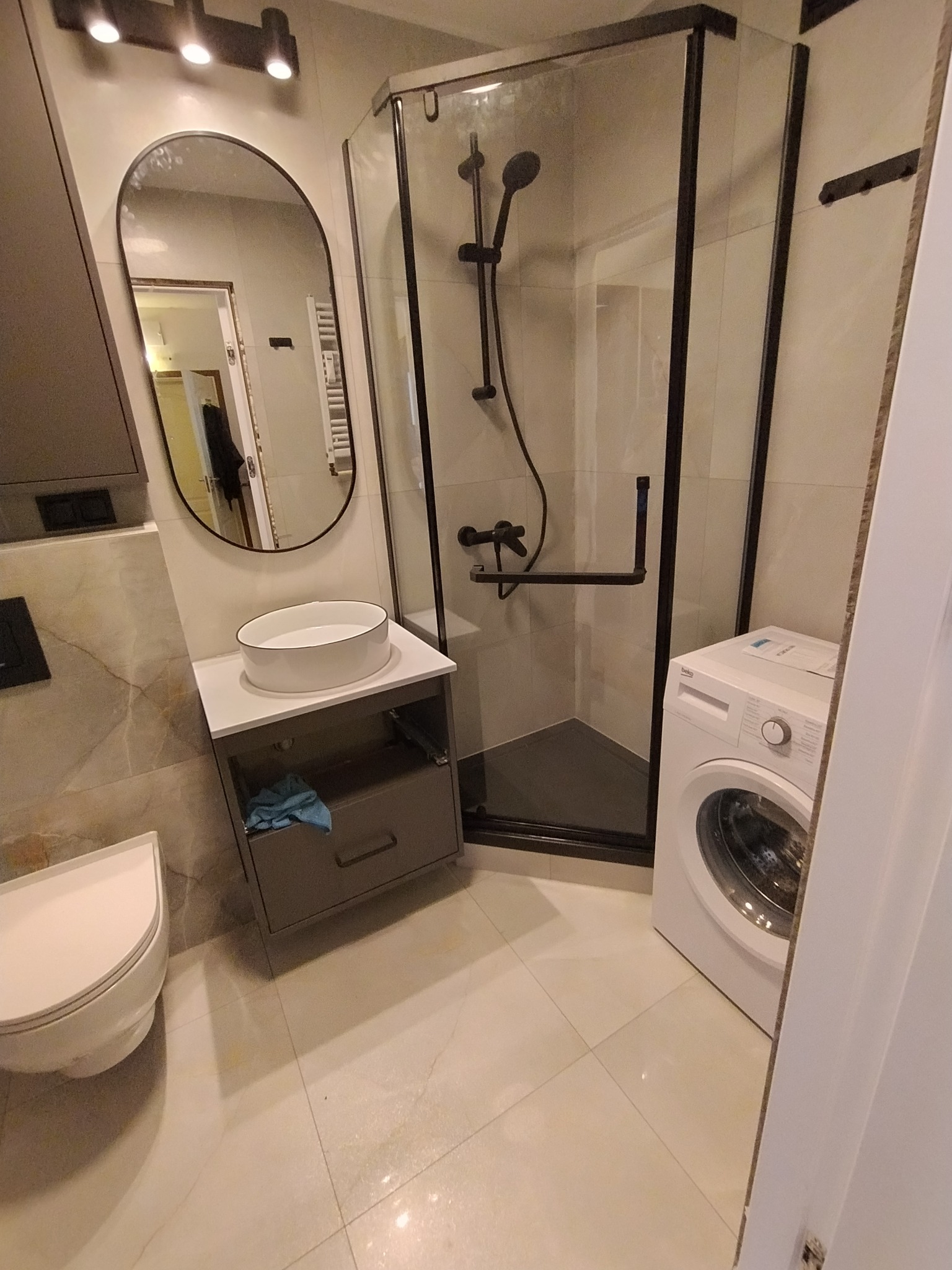 Nowoczesna łazienka z kabiną prysznicową typu walk-in z czarnymi ramami, umywalką nablatową, wiszącą szafką pod umywalką z szufladą, pralką i podświetlanym lustrem o zaokrąglonym kształcie.