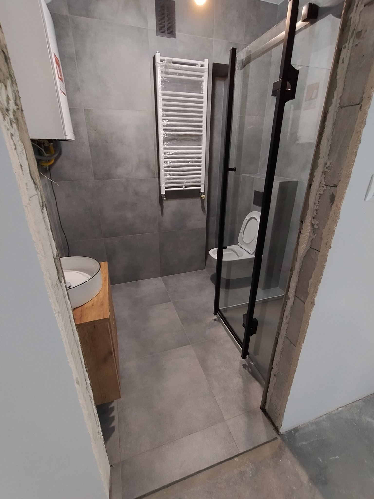 Wnętrze łazienki w trakcie remontu z widoczną kabiną prysznicową z czarnymi profilami, umywalką nablatową na drewnianej szafce, grzejnikiem drabinkowym i toaletą podwieszaną, szare płytki...