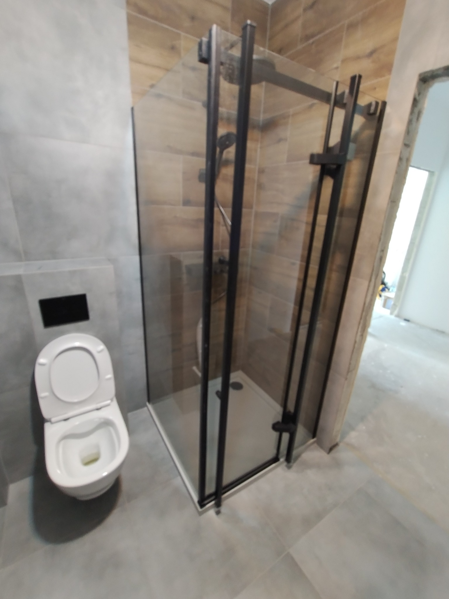 Nowoczesna łazienka z kabiną prysznicową z czarnymi profilami i wiszącą toaletą, wykończona płytkami imitującymi drewno i szarym gresem.