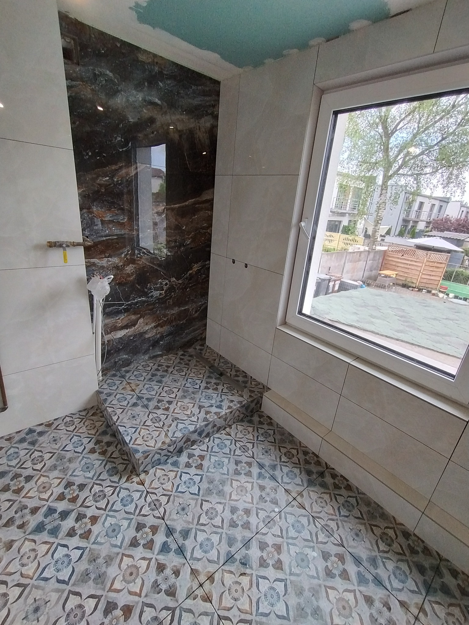 Wykończona łazienka z wzorzystą podłogą i częściowo wykończoną kabiną prysznicową z ciemnym, marmurkowym wzorem, widoczne okno z widokiem na ogród.