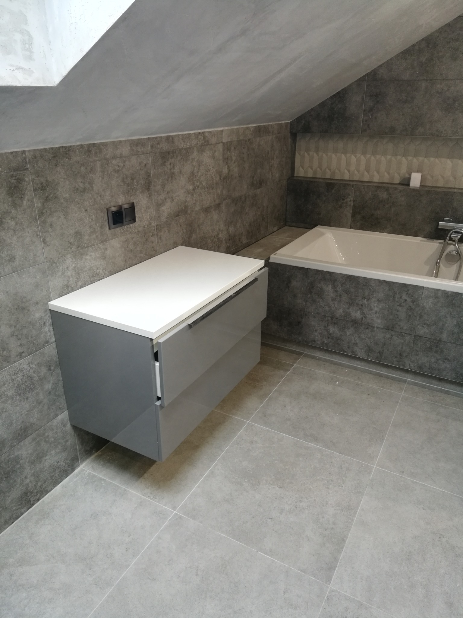 Szara łazienka z wanną i szafką pod umywalkę, płytki imitujące beton na ścianach i podłodze, skośny sufit.