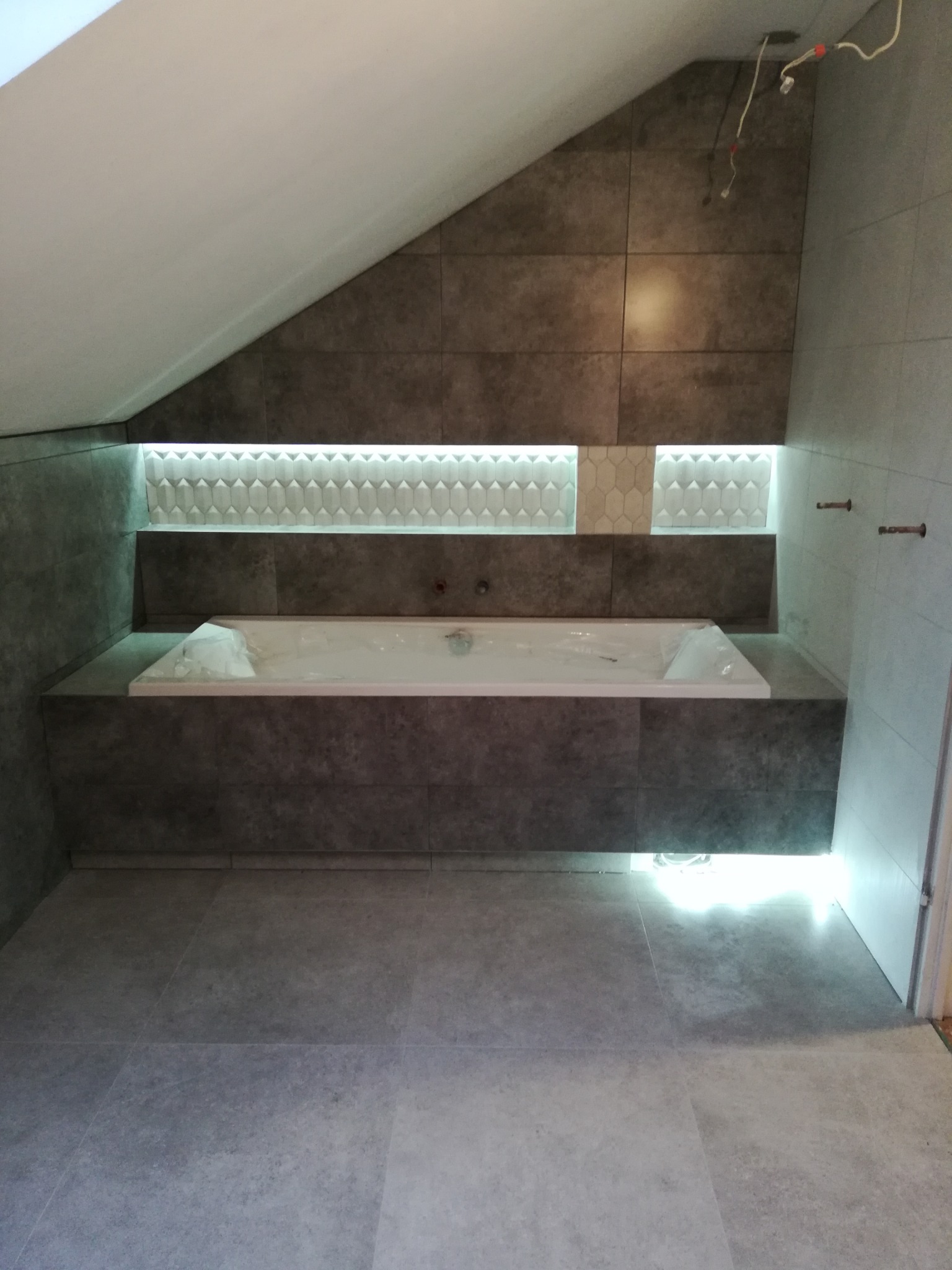 Nowoczesna łazienka w trakcie wykańczania, z obudowaną wanną i podświetleniem LED, szare płytki imitujące kamień na ścianach i podłodze, widoczne niedokończone prace elektryczne.