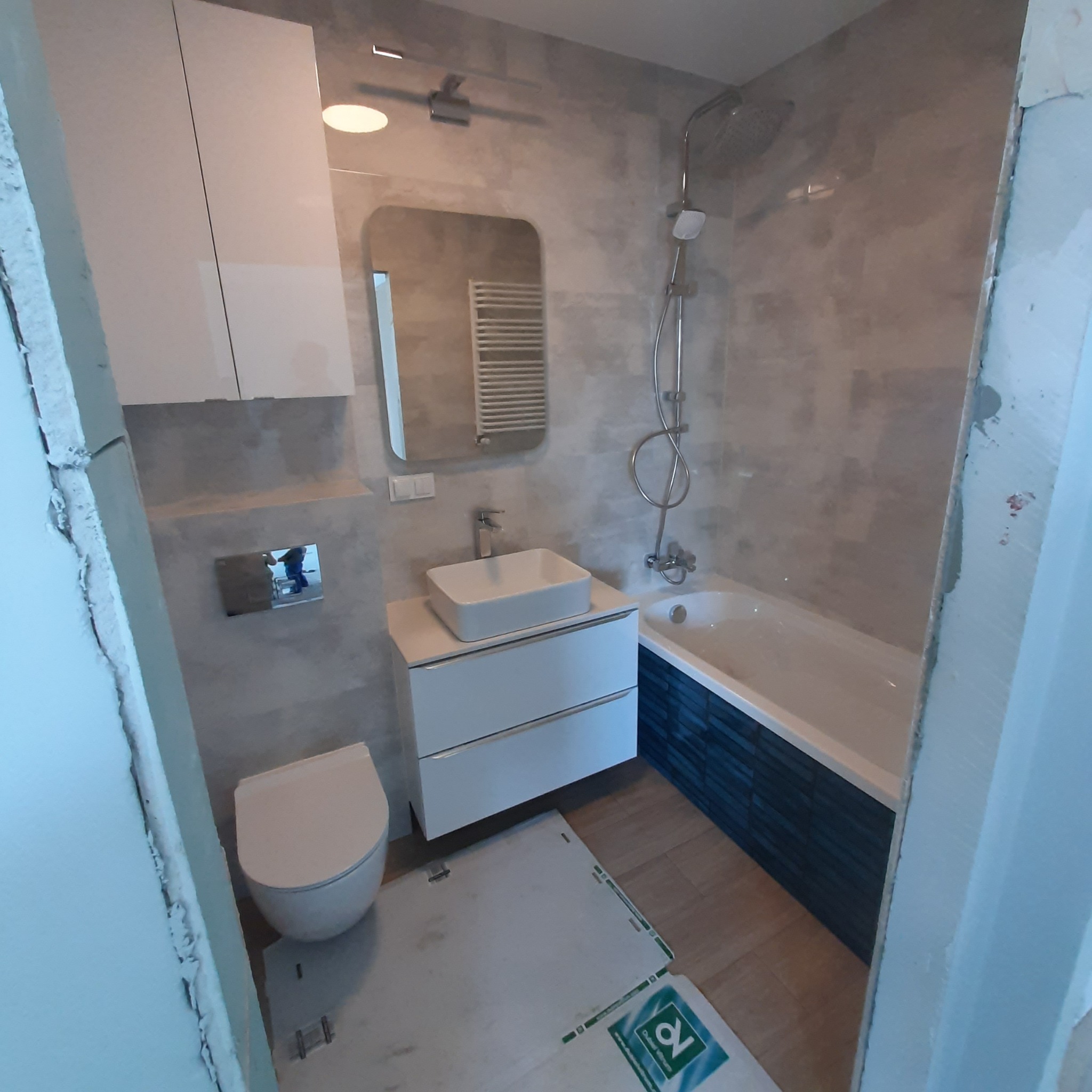 Nowoczesna łazienka z szarymi płytkami, białą armaturą i zabudową, widoczna wanna z niebieską obudową, umywalka nablatowa z szafką, toaleta, lustro i grzejnik drabinkowy.