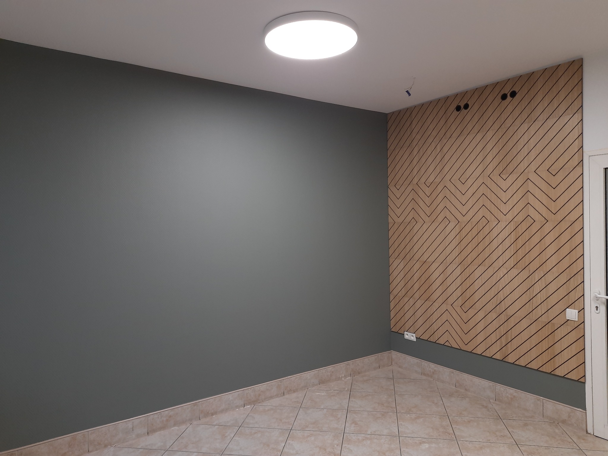 Szara ściana pomalowana farbą w pokoju z drewnianą ścianą dekoracyjną z wzorem zygzaków, jasna podłoga z płytek i okrągła lampa sufitowa.