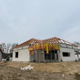 Budowa domu z białych bloczków: surowa konstrukcja, drewniana więźba dachowa z żółtymi elementami. Na pierwszym planie gruz i ziemia, w tle drugi dom i drzewa.