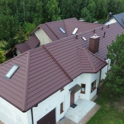 MASHOUSE Firma Budowlana - Ujęcie z lotu ptaka na dom jednorodzinny z brązowym, profilowanym pokryciem dachowym i licznymi oknami dachowymi. W tle zielony las i sąsiednie budynki.
